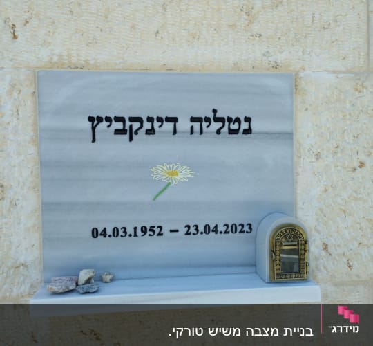 מצבה עם כיתוב בעברית, פרח ואבנים קטנות
